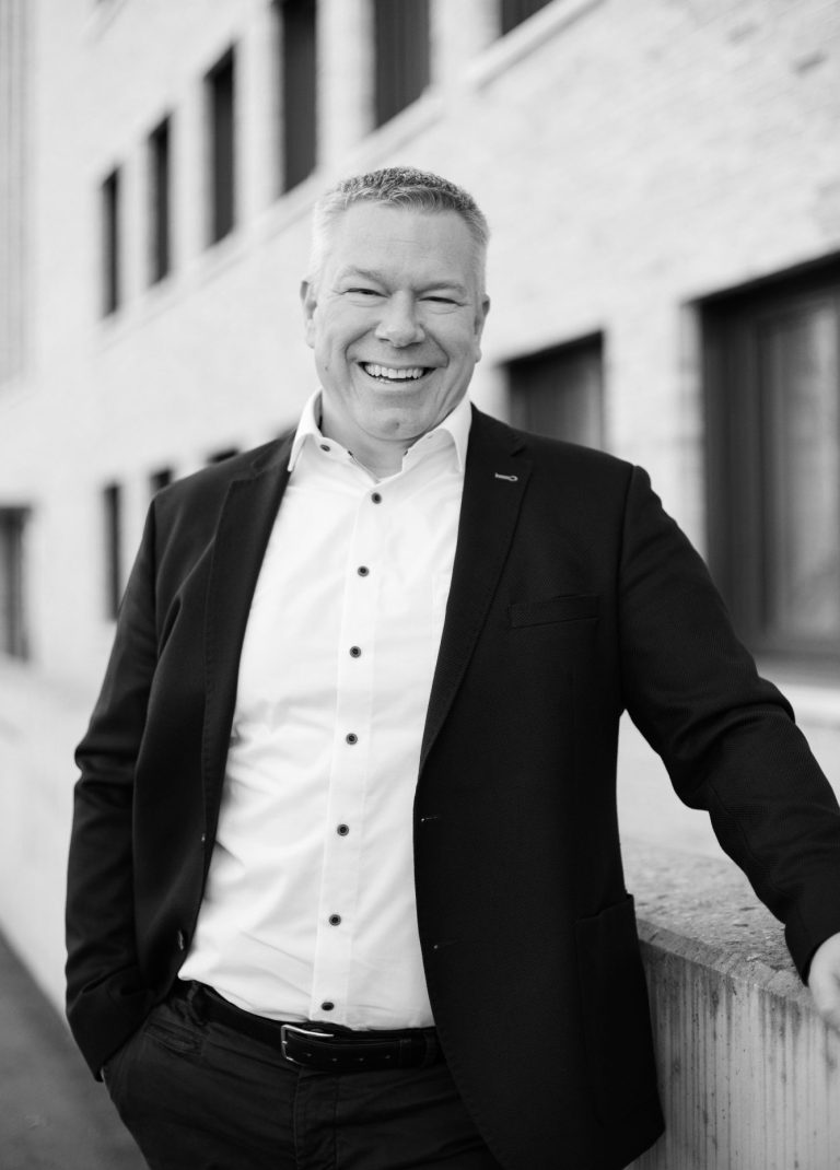 Ein Mann in den 40ern mit weißem Hemd und Jacket. Im Hintergrund ein Gebäude